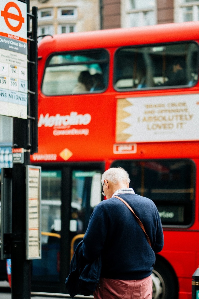 An older man waiting at a London bus stop as a red bus passes