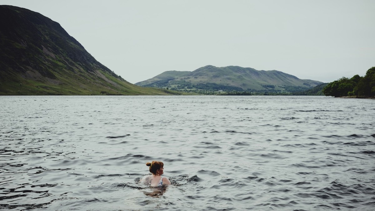 A swimmer in a still lake below green fells